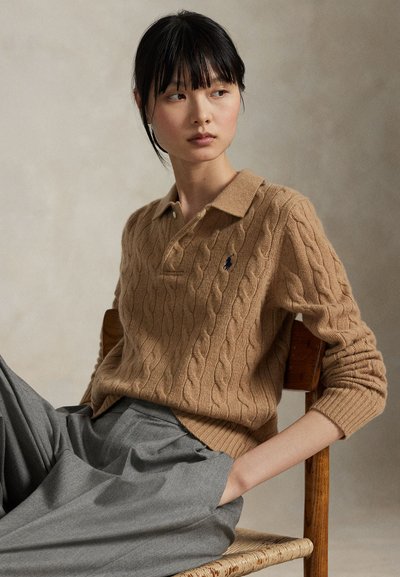 Fekete hajú nő, aki bézs színű, fonott mintás pulóvert és szürke nadrágot visel, egy fa széken ül, egyik kezét zsebében tartva, oldalra nézve.