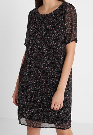 Mujer con un vestido corto de manga corta y tejido transparente en negro, con pequeños patrones florales rojos y blancos, posando frente a un fondo claro y liso.