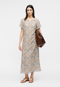 Γυναίκα που φοράει φόρεμα μήκους μέχρι τον αστράγαλο με σχέδιο πασέ με κρατά μια μεγάλη καφέ δερμάτινη τσάντα, στέκεται με σανδάλια σε απλό φόντο.