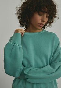 Mintgrüner übergroßer Pullover mit gerippten Bündchen und Kragen, der eine weiche Textur und einen lässigen Schnitt aufweist, teilweise sichtbar am Oberkörper einer Person.