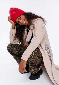 Béžový trenčkot s červenými detailmi, čierne tričko, nohavice s leopardím vzorom, červená pletená čiapka a čierne platformové topánky. Kučeravé vlasy rozpustené.