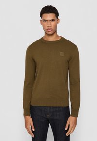 Maglione verde oliva a girocollo in un morbido tessuto a maglia, con maniche lunghe e una piccola etichetta quadrata sul petto, abbinato a jeans scuri.
