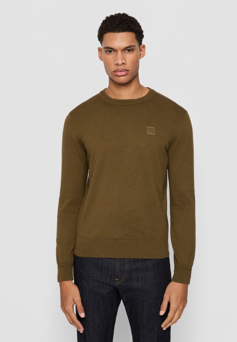 Maglione verde oliva a girocollo in un morbido tessuto a maglia, con maniche lunghe e una piccola etichetta quadrata sul petto, abbinato a jeans scuri.