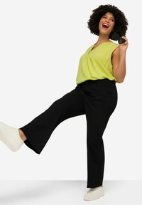 Top senza maniche di un verde chiaro con colletto, pantaloni neri a gamba larga e sneakers bianche, con una mano che tiene degli occhiali da sole neri.