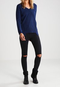 Pull en maille bleu marine à col en V, coupe ample avec fentes sur les côtés, associé à un jean skinny noir déchiré aux genoux et des bottines noires à la cheville.