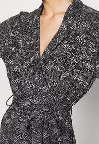 Svartvit mönstrad klänning med inslag av omslag, V-hals, korta ärmar och dragsko i midjan. Slät, lätt tyg med varierande texturer.