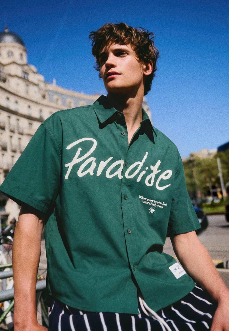 Young man wearing green button-up shirt with "Paradise" text, standing outdoors with historic building and clear blue sky in the background.