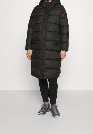 Persoon draagt een lange zwarte donsjas met capuchon, zwarte broek en zwart-grijze sportschoenen, staand tegen een effen achtergrond.