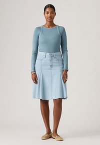 Γυναίκα στέκεται φορώντας μακρυμάνικο μπλε τοπ, φούστα μέχρι το γόνατο από ανοιχτό ντένιμ, μπεζ φλατ παπούτσια και ασημένια κοσμήματα σε απλό φόντο.