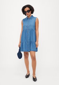 Τζιν φόρεμα με γιακά, κοντά μανίκια και κουμπιά μπροστά. Περιέχει φούστα με στρώσεις και σκούρο μπλε πλεκτή τσάντα. Μαύρες γόβες.