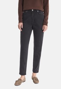 Osoba ubrana w czarne, wysokie dżinsy, brązową bluzkę z długim rękawem i beżowe mokasyny, stojąca na tle jednolitym.