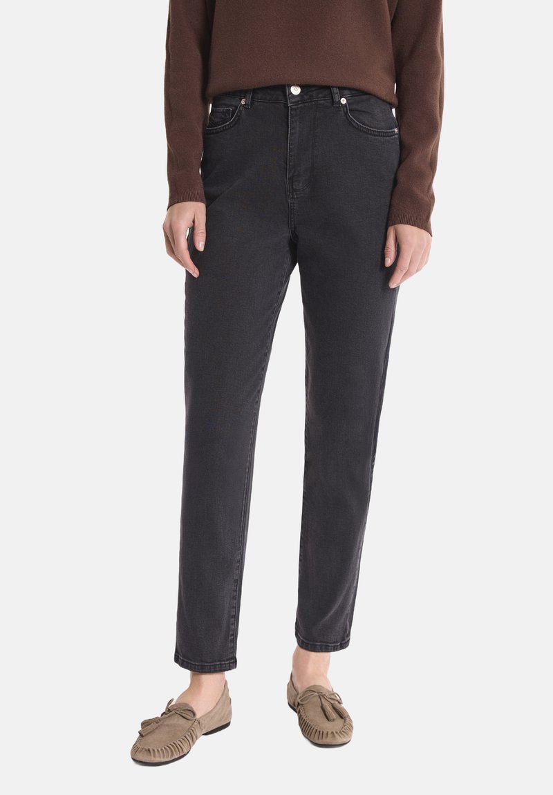 Osoba ubrana w czarne, wysokie dżinsy, brązową bluzkę z długim rękawem i beżowe mokasyny, stojąca na tle jednolitym.