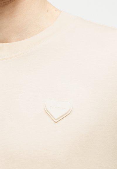 Κρεμ πουκάμισο με ένα μικρό, ραμμένο μπαλώμα σε σχήμα καρδιάς στο στήθος, που φοριέται από ένα άτομο με ανοιχτόχρωμο δέρμα.