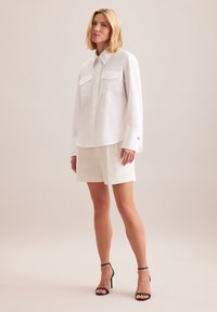 Camicia bianca a maniche lunghe con due tasche sul petto e polsini con bottone, abbinata a pantaloni corti color crema e tacchi neri a strappi, su uno sfondo neutro.
