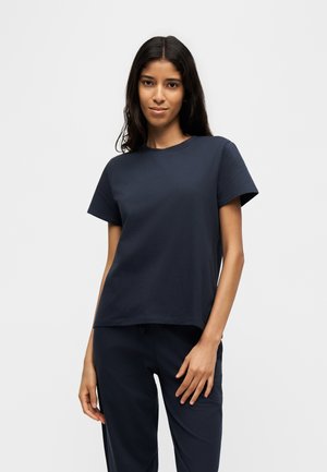 Junge Frau mit langen dunklen Haaren, die ein schlichtes dunkelblaues Kurzarmshirt und passende Hosen trägt, steht vor einem hellen Hintergrund.