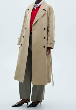 Beige dubbele-breasted trenchcoat met zwarte knopen, voorzien van brede revers en een riem in de taille; gecombineerd met olijfgroene broeken met platte voorkant en zwarte hakken.