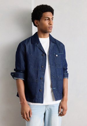 Homme portant une veste en denim bleu foncé par-dessus une chemise blanche et un jean bleu clair, debout les bras détendus, regardant à droite contre un mur uni.