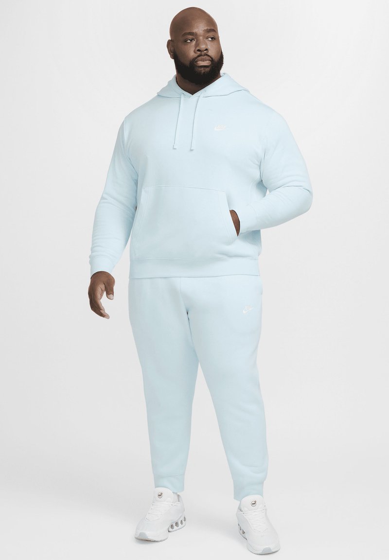 Sudadera Nike azul claro con capucha, bolsillo delantero y pantalones de chándal a juego. Logo Nike blanco en el pecho y en los pantalones. Textura de tela suave.