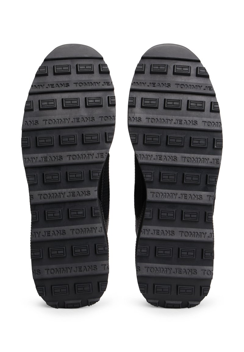 Suelas de zapatos de goma negras con un patrón texturizado que presenta formas rectangulares elevadas y la marca "TOMMY JEANS" en relieve a lo largo de la superficie.