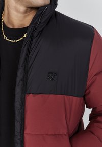 Chaqueta acolchada con una sección superior negra y una sección inferior burdeos, con un acabado texturizado y un logotipo bordado en el pecho.