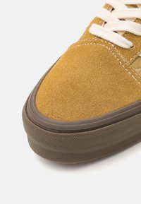 Semišové tenisky se žlutým svrškem, bílými tkaničkami a hnědou gumovou podrážkou. Zaoblená špička a strukturované strany nabízejí tradiční design skateboardových bot.