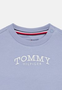 Sudadera de color púrpura claro con cuello redondo acanalado, que presenta el texto bordado "TOMMY HILFIGER" y un detalle de botones a presión cerca del hombro.