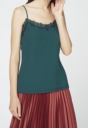 Teal camisole med blondekant ved halsudskæringen, med tynde stropper. Sammen med en plisseret, rosenfarvet nederdel. Glat tekstur.