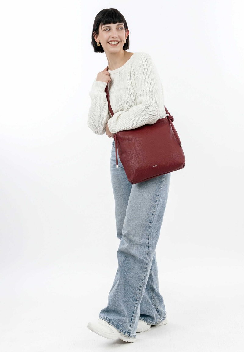 Sac à bandoulière en cuir rouge avec une surface texturée, une bandoulière ajustable et une fermeture à cordon. Forme carrée compacte avec un branding discret.