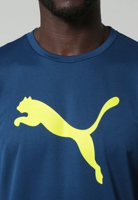 Mörkblå atletisk t-shirt av tyg med en slät textur; har en stor gul Puma-logga på bröstet.