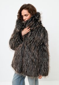 Palton din blană sintetică cu guler înalt, având fire lungi, texturate în nuanțe de negru și maro. Designul este supradimensionat, cu o croială lejeră.