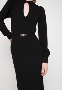 Robe noire à manches longues avec encolure goutte d'eau et ceinture fine ornée d'une boucle double anneau dorée, portée par une personne sur un fond uni.