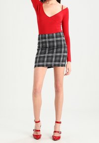 Roter langärmeliger, taillierter Body, kombiniert mit einem schwarz-weißen karierten Minirock. Das Modell trägt rote Riemchensandalen. Glatter Stoff und maßgeschneiderte Passform.