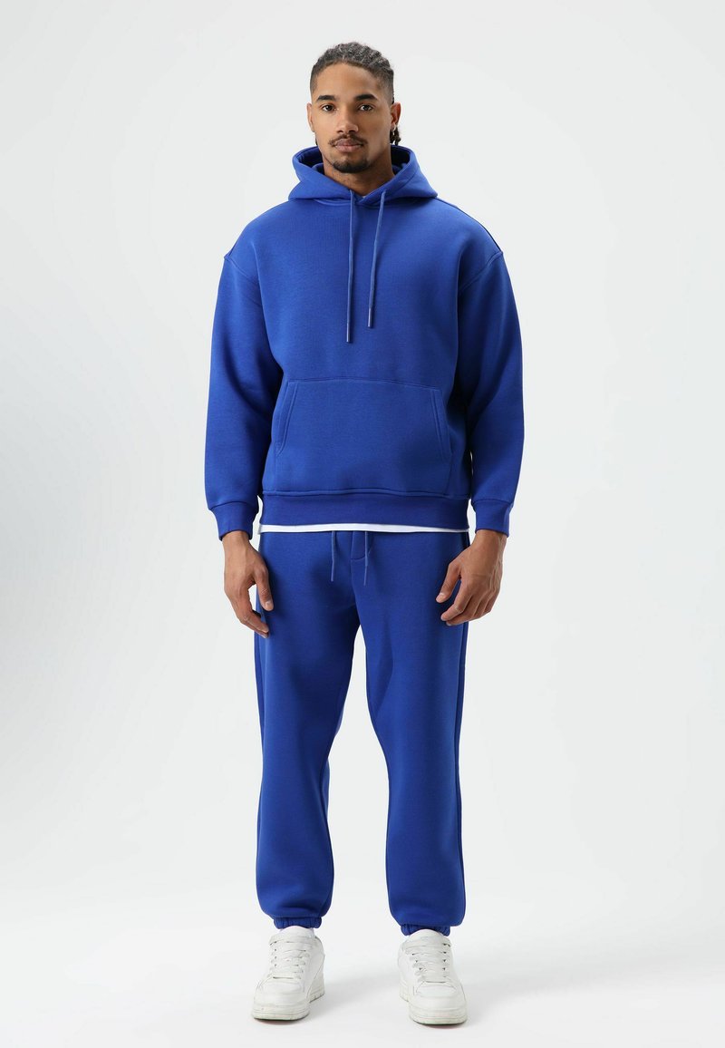 Blue hooded sweatshirt and matching joggers made of soft, textured fabric. Features a front pocket and white drawstrings, paired with white sneakers.