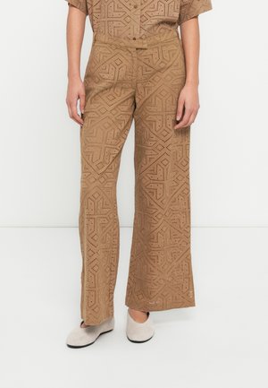 Pantalon marron à jambes larges en tissu texturé avec des motifs géométriques perforés. Associé à des chaussures beige clair.