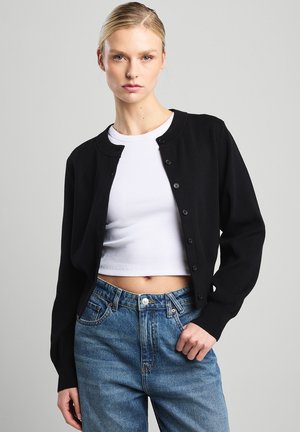 Schwarze Strickjacke mit rundem Ausschnitt und Knopfverschluss. Strickstruktur, lange Ärmel, kombiniert mit einem weißen Cropped-Top und blauen Jeans.
