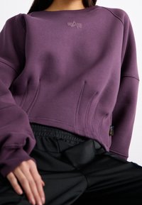 Fialová krácená mikina s kulatým výstřihem, texturovanými panely a malým logem na hrudi; v kombinaci s černými kalhotami s elastickým pasem.