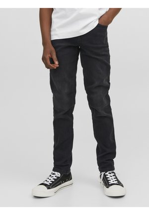 Personne portant un jean noir, un t-shirt blanc et des baskets basses noires avec des lacets blancs, debout devant un fond gris uni.