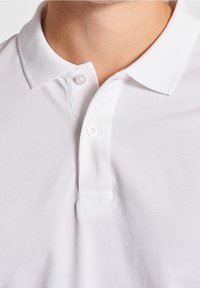 Detailní záběr na osobu v bílém polo tričku s dvěma bílými knoflíky zapnutými pod límcem.