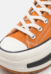 Oranžové plátěné tenisky s bílými tkaničkami, gumovou špičkou a černými akcenty podél podrážky. Obsahují detaily šití a průvleky.