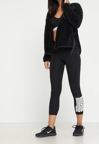 Zwarte pluizige jas met een strik aan de voorkant, gecombineerd met zwarte leggings met een witte "JUST"-logo op het onderbeen, en zwarte sportieve schoenen.