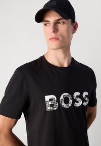 Schwarzes T-Shirt mit einem strukturierten weißen "BOSS" Grafikdruck. Das Modell trägt eine schwarze Mütze. Glatter Baumwollstoff, enganliegendes Design.