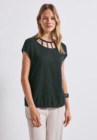 Top verde scuro a maniche corte con scollatura rotonda e dettagli con ritagli. Tessuto morbido, vestibilità ampia e orlo che si ferma sui fianchi.