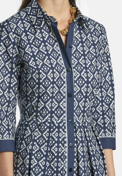Frau in einem knielangen Hemdblusenkleid mit marineblau-weißem Muster, 3/4-Ärmeln und einer goldenen Halskette.