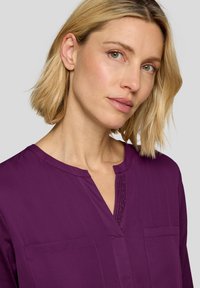 Lila Bluse mit V-Ausschnitt, kurzen Ärmeln und einer dekorativen Reihe von Nieten entlang der Knopfleiste. Glatter Stoff mit zwei Fronttaschen.