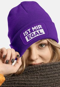 Purple knit beanie features a rectangular white patch with the text "IST MIR EGAL." The material appears soft and textured, fitting snugly on the head.