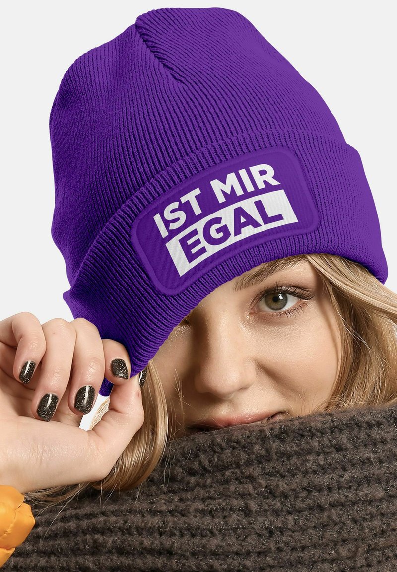 Purple knit beanie features a rectangular white patch with the text "IST MIR EGAL." The material appears soft and textured, fitting snugly on the head.