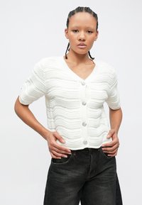 Λευκό πλεκτό ζακέτα με κυματιστά σχέδια, κοντά μανίκια και διακοσμητικά κουμπιά. Συνδυάζεται με μαύρο τζιν παντελόνι.