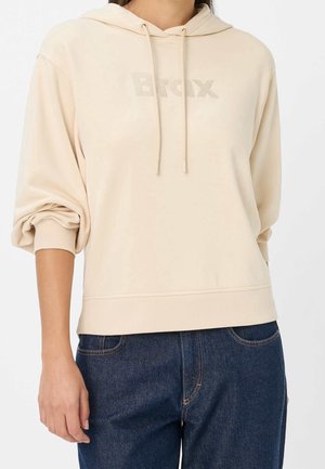 Personne portant un sweat à capuche beige avec les manches retroussées et un logo "Brax" discret, associé à un jean en denim bleu foncé.
