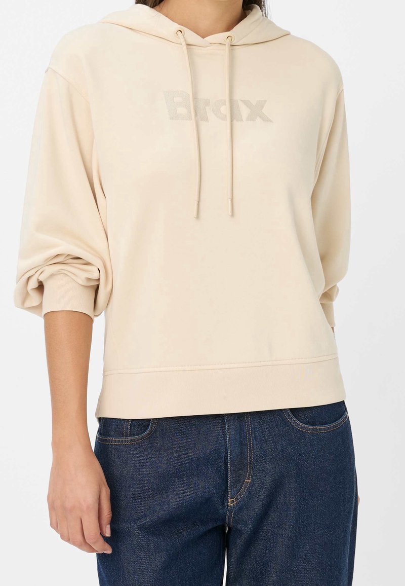 Personne portant un sweat à capuche beige avec les manches retroussées et un logo "Brax" discret, associé à un jean en denim bleu foncé.