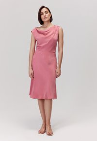 Różowa sukienka z dekoltem typu cowl, krótkimi rękawami i sylwetką w linii A. Gładki materiał z delikatnym połyskiem, długość do kolan. Podkreślona talia.